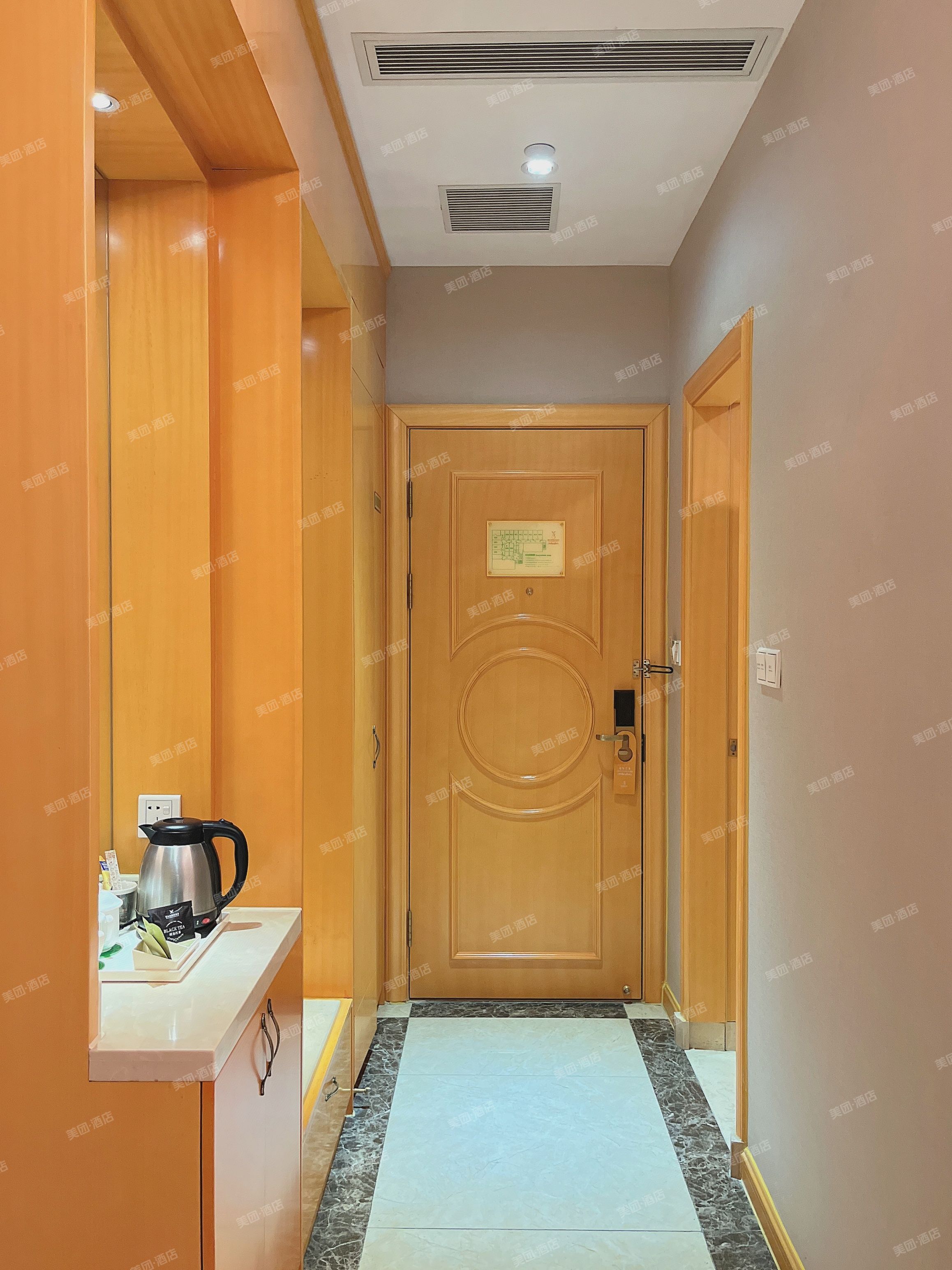 Corredor de hotel com porta de madeira, pequena cozinha com chaleira e piso ladrilhado.
