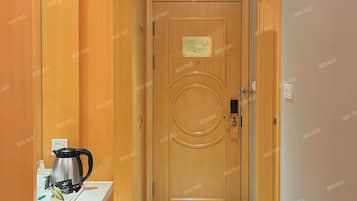 Corredor de hotel com porta de madeira, pequena cozinha com chaleira e piso ladrilhado.