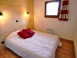 A bedroom with a wooden slat ceiling, a bed with white bedding and red pillows, a window with curtains, and a wall-mounted heater. - Spacious apartment, pets welcome - 6 guests. Park view (Villarodin-Bourget)