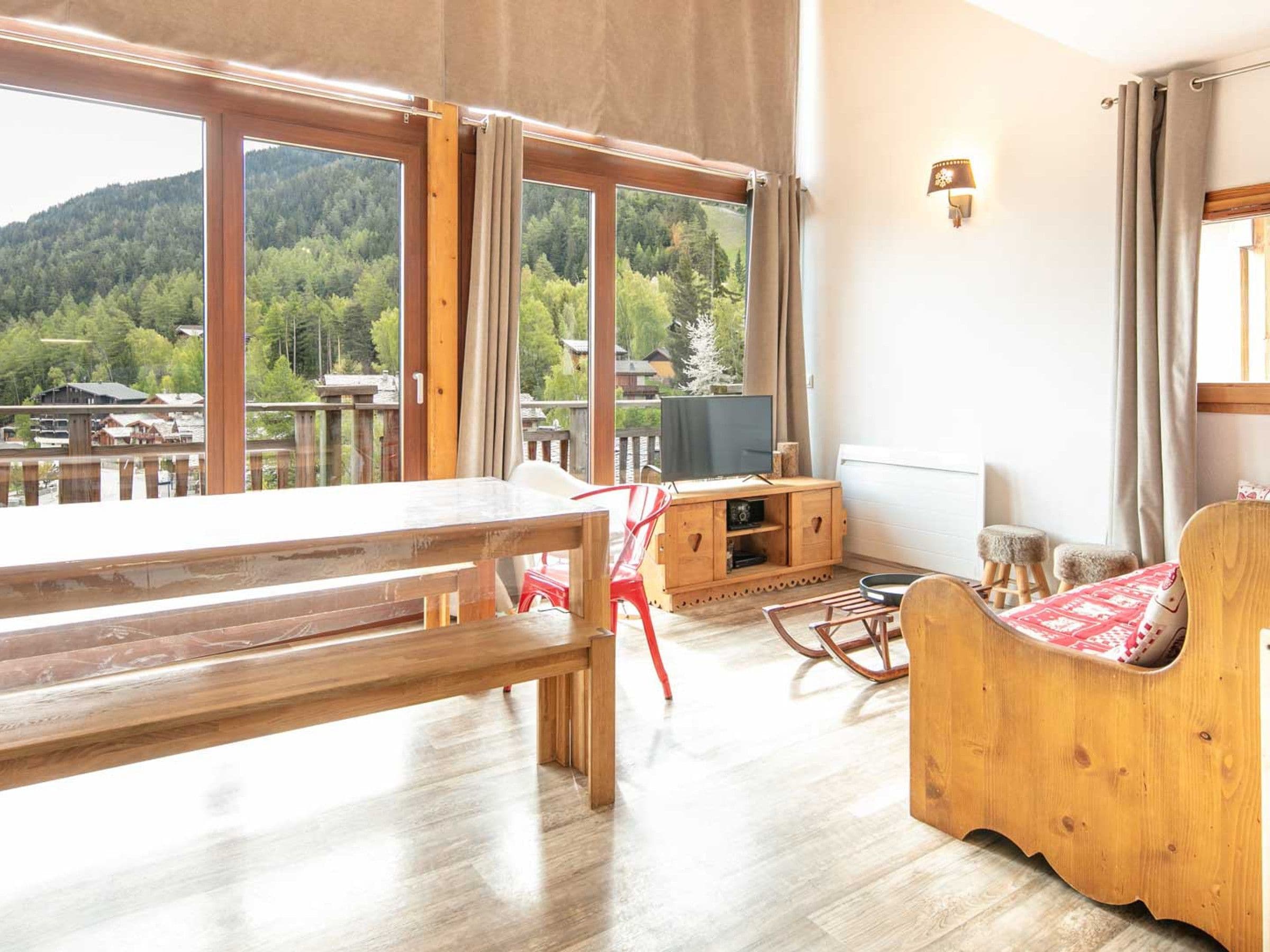 A room with a wooden table, a TV on a wooden stand, a sofa, and a view of a forested area through large windows.