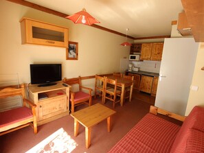 Een compact woongedeelte met een televisie, houten meubels, een eettafel en een keukenhoek.