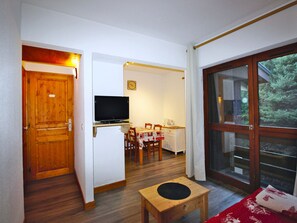 A compact living space with a wooden door, a television on a stand, a dining area with a table and chairs, and a large window offering a view of greenery. - Cozy and quiet studio - 4 guests (Villarodin-Bourget)