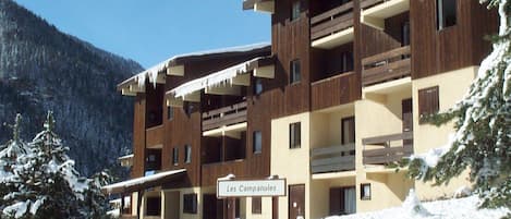 A multi-story building with balconies, a sign indicating 'Les Campanules,' and a snowy landscape with trees.