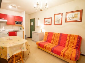 A compact living space with a kitchenette, a dining table with a floral tablecloth, a sofa with a patterned blanket, and three framed pictures on the wall. - Quiet apartment by the forest - 4 guests (Villarodin-Bourget)