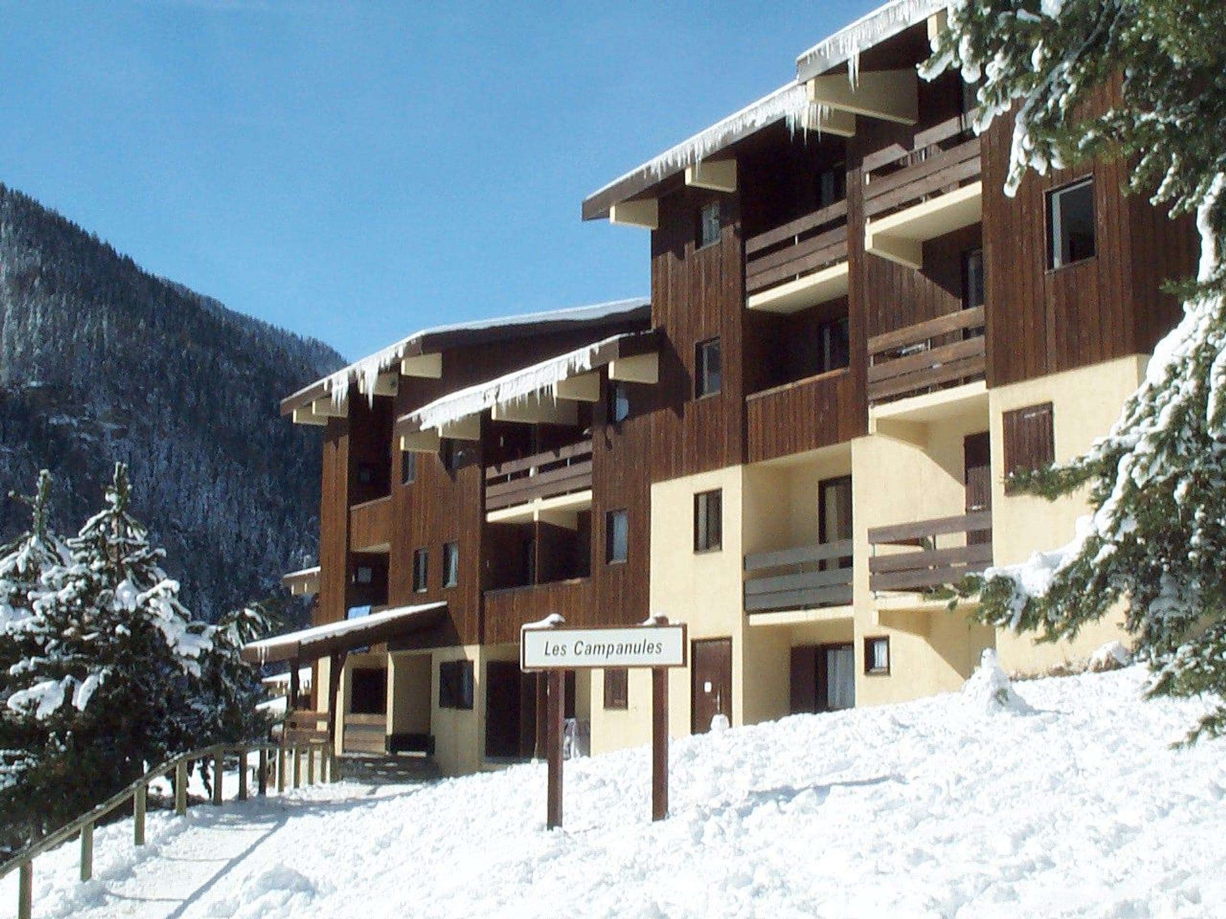 A multi-story building with balconies, a sign indicating 'Les Campanules,' and a snowy landscape with trees.
