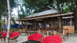 An outdoor seating area with wicker chairs and tables, a rustic wooden building with a corrugated metal roof, and a sign indicating a restaurant. - Rock Sand Hostel (Koh Rong)