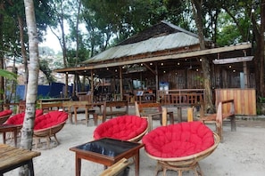 An outdoor seating area with wicker chairs and tables, a rustic wooden building with a corrugated metal roof, and a sign indicating a restaurant. - Rock Sand Hostel (Koh Rong)