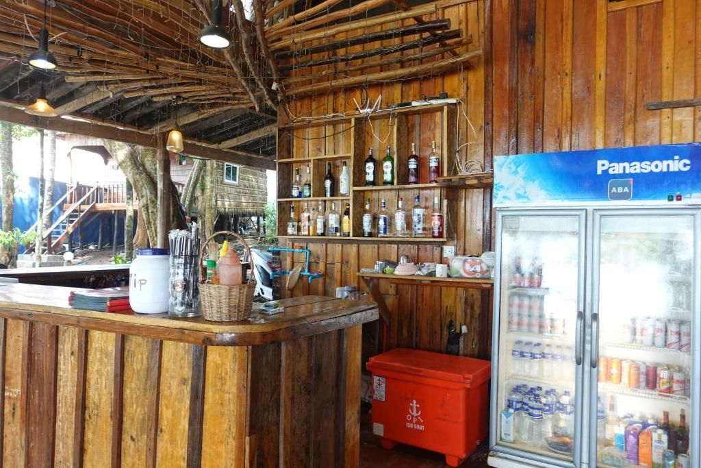 Uma área de bar com balcão de madeira, geladeira repleta de bebidas, bancada com diversos itens e prateleira com garrafas.