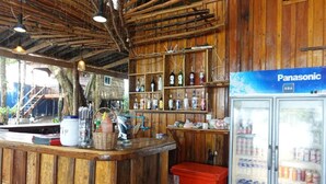 A wooden bar area with a refrigerator stocked with beverages, a counter with various items, and a shelf with bottles.