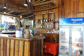 A wooden bar area with a refrigerator stocked with beverages, a counter with various items, and a shelf with bottles.