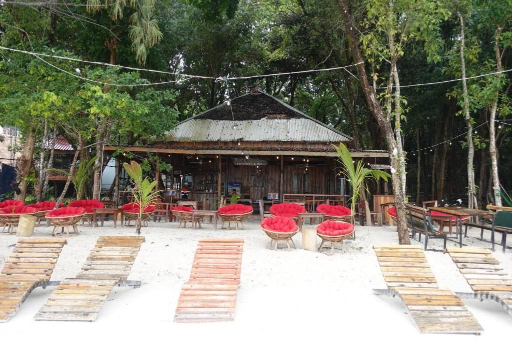 Un edificio rústico de madera con techo de metal corrugado, rodeado de sillas con cojines rojos y mesas de madera, con abundante vegetación de fondo.