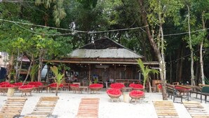 A rustic wooden building with a corrugated metal roof, surrounded by red cushioned chairs and wooden tables, set against a backdrop of dense greenery.