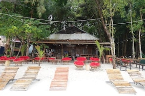 A rustic wooden building with a corrugated metal roof, surrounded by red cushioned chairs and wooden tables, set against a backdrop of dense greenery.