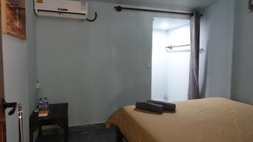 A hotel room with a bed, an air conditioning unit, a small table with bottled water, and a door leading to another room.
