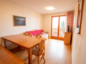 Una habitación con una mesa de comedor de madera, sillas, un sofá con una manta roja, un cuadro en la pared y un ventanal corredero que da a un espacio exterior.