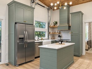 A modern kitchen with a stainless steel refrigerator, a central island, and a window overlooking greenery. - Lakehaus at Water Edge -Modern Luxury/Beach/Dock/Kayaks/Fire Pit (Tracy City)