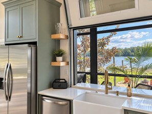 A modern kitchen with a stainless steel refrigerator, a white countertop, a sink with a gold faucet, and a view of a lake through large windows. - Lakehaus at Water Edge -Modern Luxury/Beach/Dock/Kayaks/Fire Pit (Tracy City)