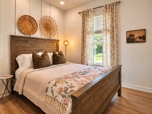 A bedroom with a wooden headboard, a floral bedspread, a bedside table, and a window with curtains. - Lakehaus at Water Edge -Modern Luxury/Beach/Dock/Kayaks/Fire Pit (Tracy City)