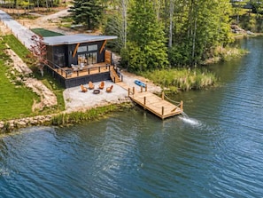 A lakeside cabin with a dock extending into the water, a deck with outdoor seating, and a forested backdrop. - Lakehaus at Water Edge -Modern Luxury/Beach/Dock/Kayaks/Fire Pit (Tracy City)