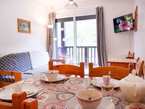 Ein Essbereich mit einem für vier Personen gedeckten Tisch, einem an der Wand befestigten Fernseher und einem Balkon mit Blick auf Grünflächen.