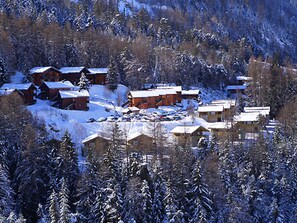 Ein verschneites Dorf mit mehreren Holzbauten, wahrscheinlich Chalets oder Ferienhäusern, umgeben von dichtem Wald.