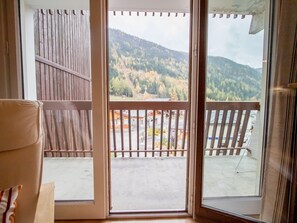 Ein Zimmer mit einer Schiebeglasdoor zur Außenbalkons, mit Holzgeländer, Blick auf einen bewaldeten Hang und eine gepflasterte Fläche mit Gebäuden im Hintergrund.