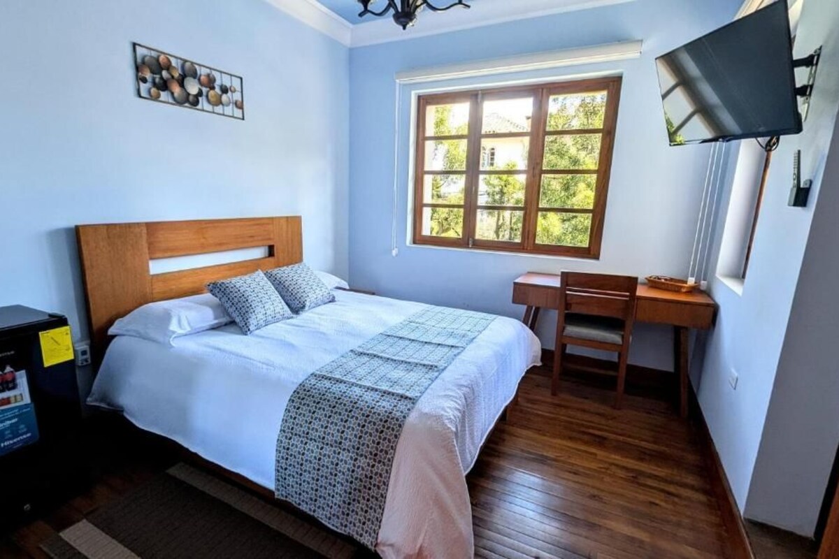 Ein Schlafzimmer mit Bett, Schreibtisch, Fernseher und Fenster mit Blick auf Grünflächen.