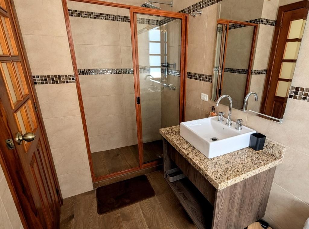 Un baño con ducha envuelta en vidrio, un lavamanos con encimera de granito y una puerta de madera.