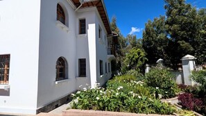Un bâtiment blanc doté de nombreuses fenêtres et d’un balcon, entouré d’un jardin où poussent diverses plantes et fleurs.