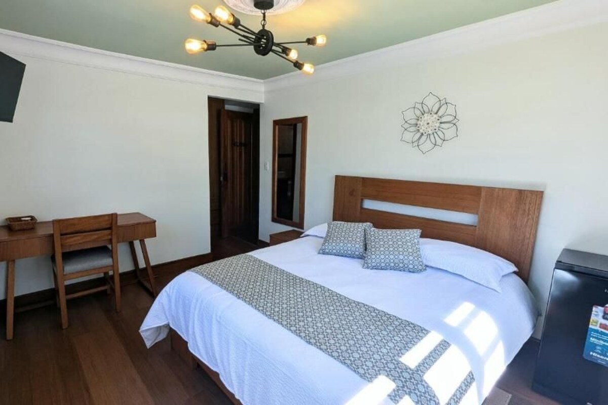 Ein Schlafzimmer mit Bett, Nachttisch, Schreibtisch, Stuhl, Fernseher und einer Deckenleuchte.