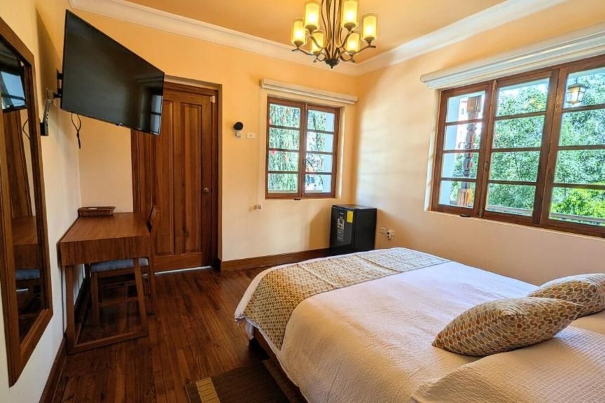 Ein Schlafzimmer mit Bett, Schreibtisch, Fernseher und Fenstern mit Blick auf Grünflächen.
