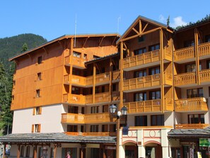 A multi-story wooden building with balconies, a shop at the ground level, and a mountain in the background. - Studio with massif view - 3 guests (Valfréjus)