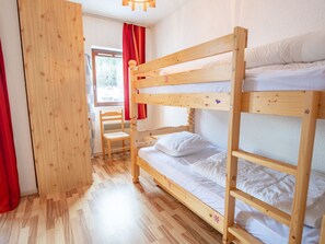 Habitació amb literes, sòl de fusta, una cadira i una finestra amb cortines vermelles.