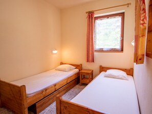 Ein kleiner, gemütlicher Raum mit zwei Holzbettstegen, einem Tisch und einem Fenster mit Vorhängen.
