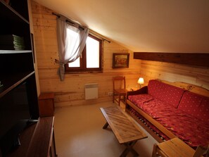 A cozy room with a wooden sloped ceiling, a single bed with a red patterned bedspread, a wooden bench, a small table with a lamp, a chair, and a wall-mounted heater. - Spacious duplex mountain view - 12 guests (Valfréjus)