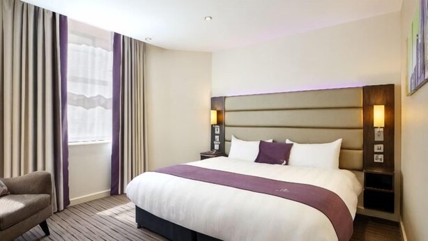 A hotel room with a large bed, a chair, a nightstand, and a window with curtains. - Milton Keynes Sw(Furzton Lake) (Furzton)