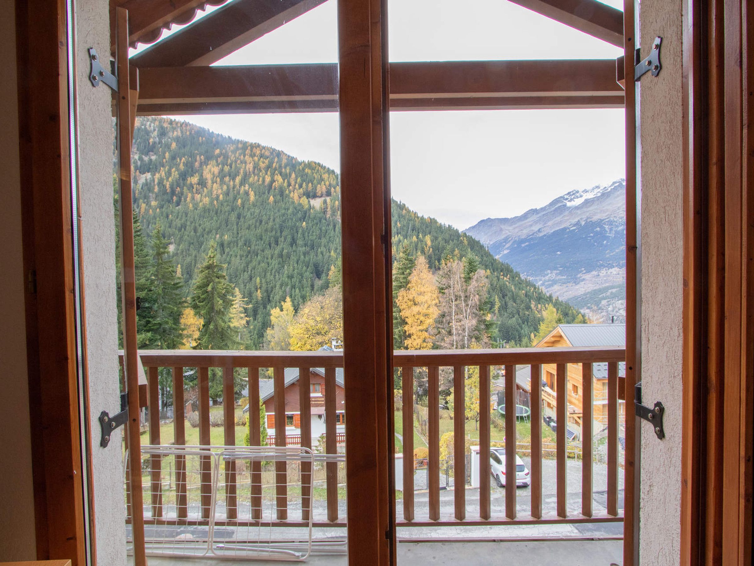 A wooden balcony with a metal railing, overlooking a mountainous landscape with autumn foliage and a residential area below.