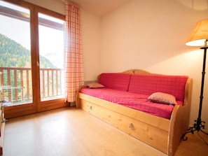 A wooden bed with a red bedspread and two pillows, a wooden floor, a window with a checkered curtain, and a lamp on the right. - Charming apartment, mountain view - 6 guests (Valfréjus)