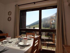 A dining area with a table set for two, a window with a view of a mountain landscape, and a wooden chair. - 2-room apartment with balcony - 4 guests (Villarodin-Bourget)