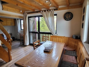 Ein Holztisch mit einer Plastikabdeckung, eine Bank mit Kissen, eine Uhr an der Wand und Blick ins Grüne.