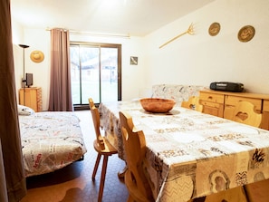 Ein Zimmer mit Esstisch, Stühlen, einem Holzschrank, einem Fernseher auf einem Schrank und einem großen Fenster mit Vorhängen.