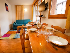 A dining area with a wooden table set for four, surrounded by chairs, with a blue sofa and a colorful rug in the background. - Apartment pets welcome - 6 guests, mountain view (Villarodin-Bourget)