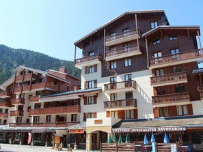 Un hotel de varios pisos con balcones, un restaurante llamado 'CREPERIE SPECIALITES SAVOYARDES' y un cartel que indica 'LOCATION DE SKIS'.