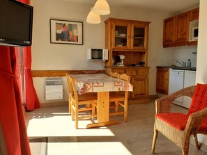 Cocina compacta con armarios de madera, una mesa de comedor con mantel a cuadros, una silla de mimbre y un televisor en la pared.