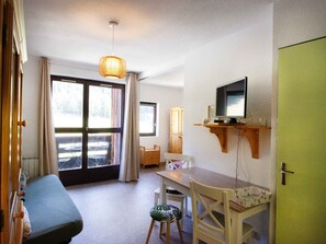 A room with a sofa, a dining table, a TV on a shelf, and a window with curtains. - Apartment with mountain view - 4 guests (Villarodin-Bourget)