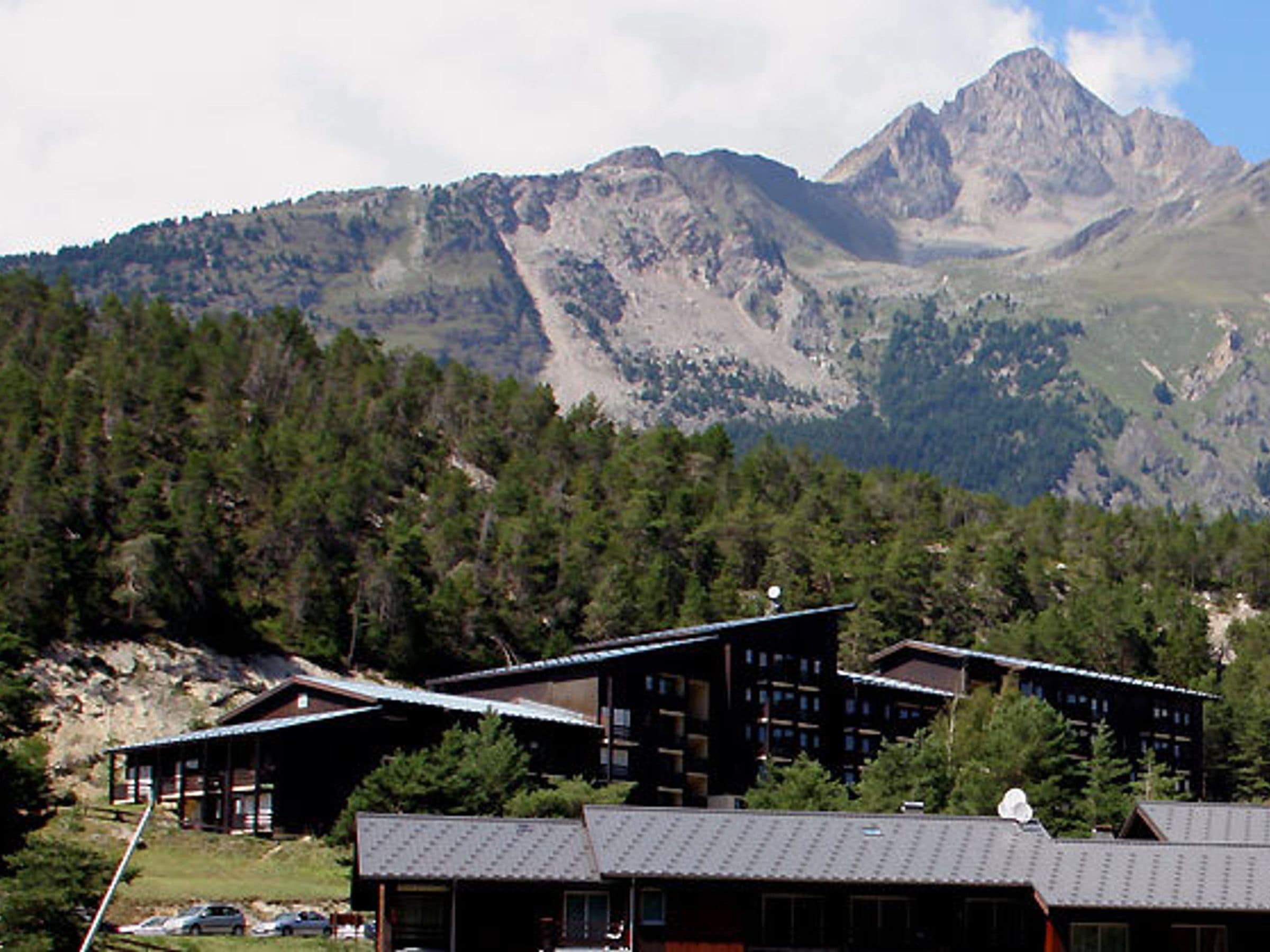 A mountain lodge with multiple buildings, surrounded by forested hills and a prominent mountain peak.