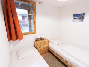 Ein kleines, sauberes Zimmer mit zwei Einzelbetten, einem Nachttisch aus Holz mit zwei Lampen, einem Fenster mit Vorhängen und einem Bild an der Wand.