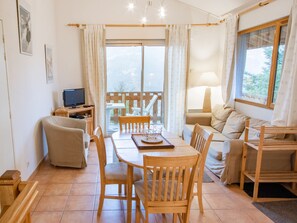 Ein gemütliches Wohnzimmer mit einem Holzesstisch, Stühlen, einer Couch, einem Fernseher und einem Balkon mit Gartenmöbeln.
