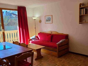 A room with a wooden sofa, a table, and a chair. There is a window with a view of a building outside. - Lovely apartment - 6 guests, near ski lift (Valfréjus)