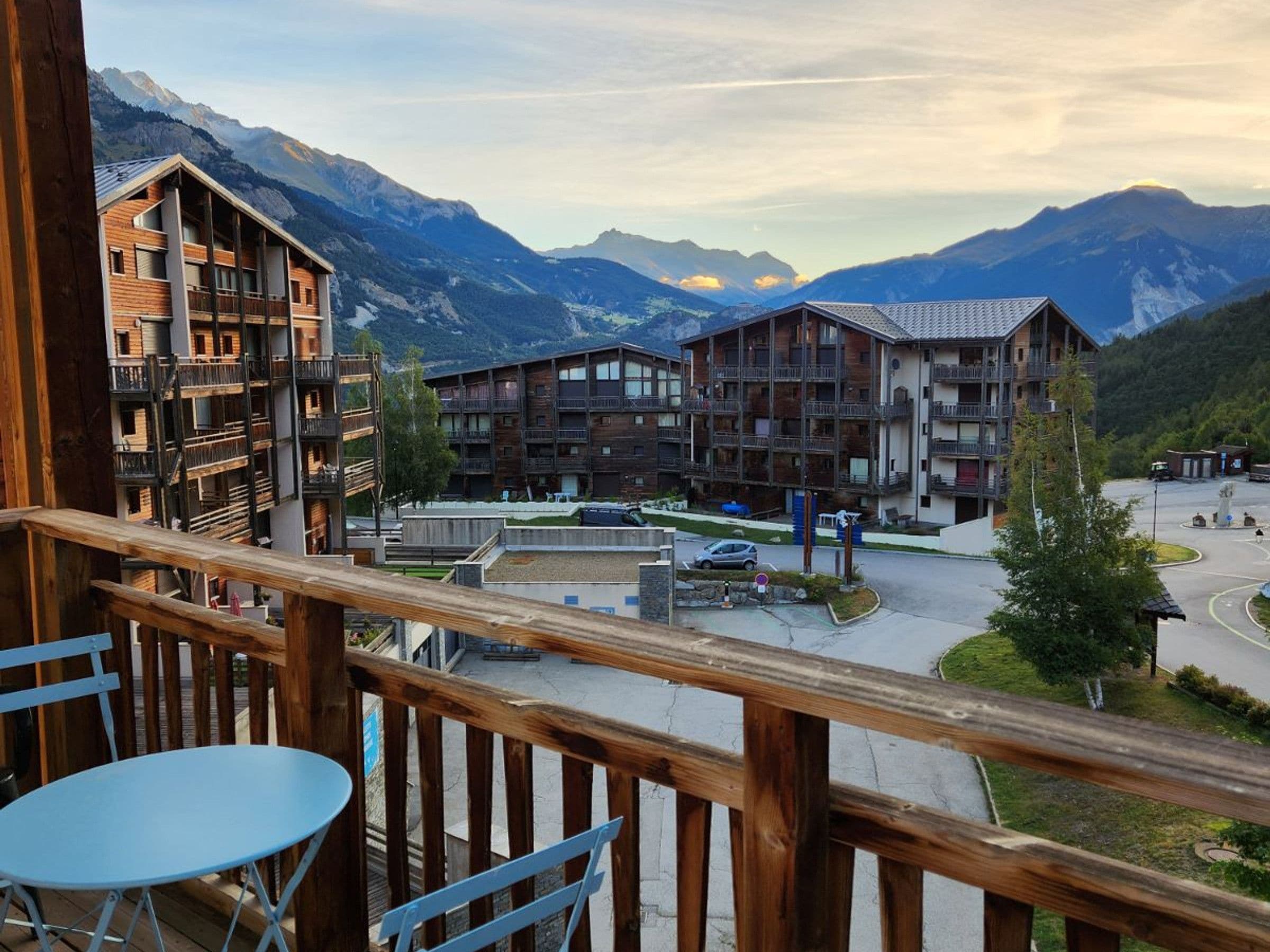 A wooden balcony with a table and chairs, overlooking a mountain resort with multiple buildings and a parking area.